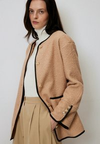 Giacca di tessuto pesca con rifiniture nere, caratterizzata da bottoni dorati e tasche laterali. Indossata sopra un dolcevita bianco e pantaloni beige a pieghe.