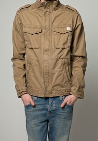 Brązowa kurtka na suwak z dwoma kieszeniami na piersi i pagonami na ramionach. Tkanina wydaje się lekka i gładka w dotyku. Noszona z jasnoniebieskimi spodniami jeansowymi.