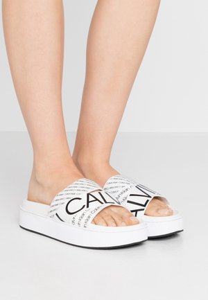 Witte slidesandalen met een dikke plateauzool en brede banden met herhaaldelijk zwart merktext. Gladde textuur met een openteenontwerp.