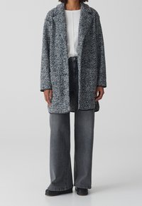 Abrigo de punto texturizado en negro y blanco, de mangas largas, con abertura frontal, combinado con jeans grises de pierna ancha de cintura alta y zapatos negros.