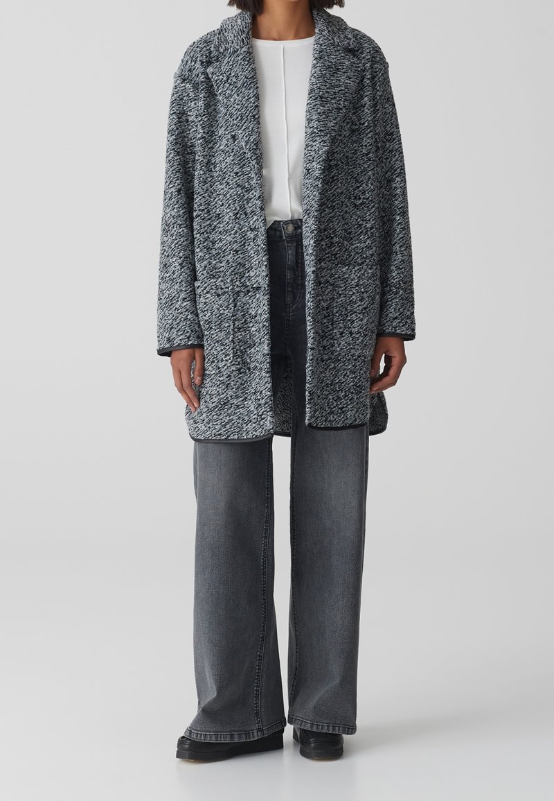 Texturerad stickad kappa i svart och vitt, lång ärm, öppen front, parat med högmidjade grå vida byxor och svarta skor.