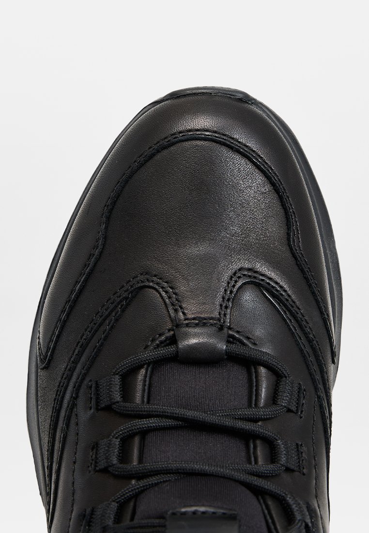 Schwarzer Leder-Sneaker mit abgerundeter Spitze, detaillierter Naht und schwarzen Schnürsenkeln auf einem schlichten hellgrauen Hintergrund.