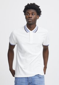 Weißes Poloshirt mit einem navy- und weißen gestreiften Kragen, kurzen Ärmeln und einem dezenten Logo auf der Brust; kombiniert mit blauen Jeans.