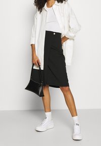 Black skirt with a front button detail, paired with a white fitted top and an oversized white button-up shirt. Worn with white sneakers.