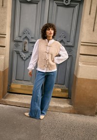 Béžová pletená vesta s textúrovanými detailmi a prednými zaväzovacími pásikmi, vrstvená cez bielu blúzku so čipkovými manžetami, v kombinácii so širokými modrými džínsami a béžovými topánkami.