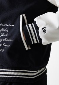 Black and white varsity jacket featuring leather sleeves, striped ribbed cuffs, and embroidered text detailing on the body.