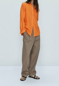 Chemise orange à manches longues, associée à un pantalon beige ample et des sandales noires. Tissu lisse et coupe décontractée.