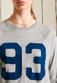 Harmaa collegepaita, jossa on tummansinisesti painettu numero "93", pyöreä pääntie ja raglanhihoja. Kulta kaulaketju, jossa on pieniä yksityiskohtia.