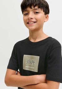 Zwart katoenen T-shirt met korte mouwen, voorzien van een rechthoekige afbeelding in roomkleur en goud met de tekst "JACK & JONES" in vetgedrukte letters.