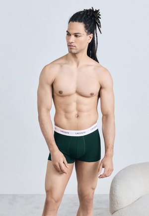 Άνδρας χωρίς πουκάμισο με ντρέντλοκς, φορώντας σκούρα πράσινα μποξέρ με λευκή ζώνη μέσης Lacoste, στέκεται κοντά σε μια λευκή υφασμάτινη καρέκλα.