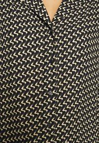 Schwarz-cremefarbene Hemdbluse mit Zickzackmuster, Kragen und fünf Knöpfen vorne. Der Stoff wirkt leicht und strukturiert.