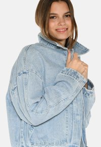Svijetloplava traper jakna s visokim ovratnikom, oversize kroja i vidljivim šavovima. Ima velike džepove i zatvaranje na patentni zatvarač.