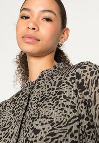 Blusa transparente e estampada em verde oliva suave com manchas abstratas pretas. Apresenta um decote redondo e detalhe em botão. Textura suave e leve.