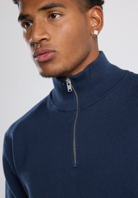 Navy blue zip-neck sweater made of ribbed fabric. Features a high collar and a silver zipper, showcasing a textured, fitted design.