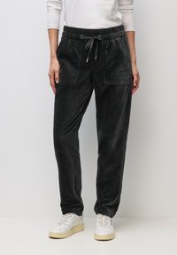 Donna che indossa pantaloni di velluto a coste neri con coulisse e tasche frontali, e sneakers bianche, in piedi davanti a uno sfondo chiaro uniforme.