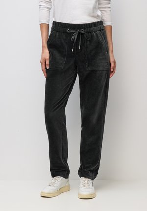 Nainen pukeutuneena mustiin samettinauhakappaleisiin, joissa on etutaskut, ja valkoisiin tennareihin, seisomassa yksinkertaisen vaalean taustan edessä.