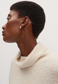 Maglione lavorato a maglia di un caldo color crema chiaro con collo a imbuto. Orecchino a cerchio argento visibile. Textura liscia e sottile lucentezza sotto una luce morbida.