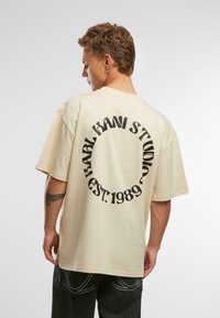 Šviesiai ruda, per didelė medvilninė marškinė, su juodu apvaliu logotipu ir inscription "BRAND KANI STUDIO EST. 1989" ant nugaros. Trumpomis rankovėmis.