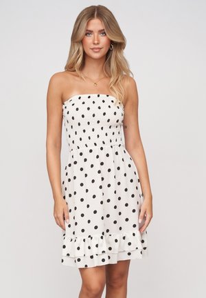 Blondine vrouw draagt een strapless witte jurk met zwarte stippen en een geplooide zoom, staand tegen een lichtgrijze achtergrond.