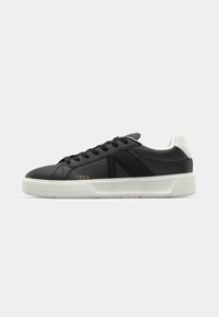 Svart skinnsneaker med hvit gummisåle. Har svarte snørebånd og et minimalistisk design. Gull ARKK-logo på siden.
