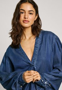 Top in denim con scollo a V profondo, ampie maniche a palloncino e polsini con bottoni. Il tessuto presenta una tonalità di blu scuro e una texture sottile.