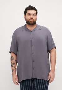 Camisa de manga corta gris en una tela ligera con un cierre de botones en la parte delantera y un corte relajado. Presenta un collar con muesca y una textura suave.