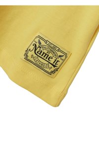 Esquina de una camiseta de algodón amarilla con una etiqueta tejida que presenta texto negro y elementos decorativos, indicando la marca "Name it Clothing Co."