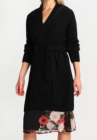 Black knitted robe with a textured design, wrap front, long sleeves, and a matching belt. Paired with a floral print dress underneath.