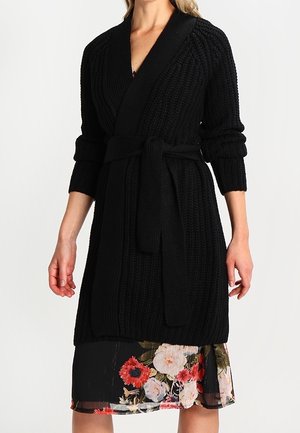 Accappatoio nero lavorato a maglia con un design testurizzato, con chiusura avvolgente, maniche lunghe e una cintura coordinata. Abbinato a un vestito con stampa floreale sotto.