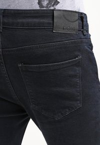 Dark denim jeans with a smooth texture, featuring a back pocket and a rectangular black label with the text "KIOMI" near the waistband.