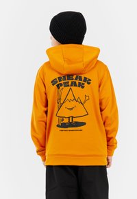 Oranžová mikina s kapucňou s čiernym grafickým dizajnom, ktorý zobrazuje horu a postavu, zobrazená zozadu. Na hlave je čierny rebrovaný čiapka.