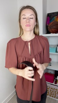 Braune Bluse mit einem Ausschnitt in Schlüssellochform, schwarze Träger in den Händen haltend, und Regale mit verschiedenen Gegenständen, darunter ein Basketball und farbenfrohe Lampen.