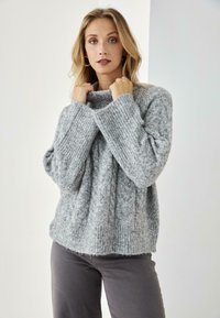 Suéter gris de punto con un patrón de cable, puños acanalados y un cuello alto. Textura suave, corte holgado, combinado con pantalones grises.