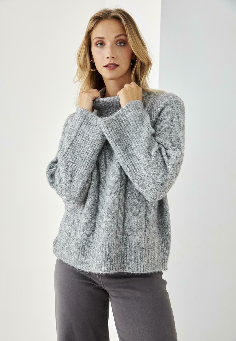 Suéter gris de punto con un patrón de cable, puños acanalados y un cuello alto. Textura suave, corte holgado, combinado con pantalones grises.