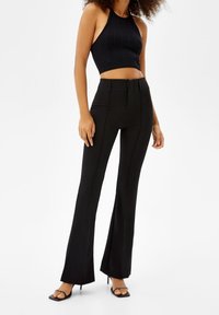 Kvinna i svart ärmlös crop top, högmidjade svarta utsvängda byxor och svarta sandaler med öppen tå mot en vit bakgrund.