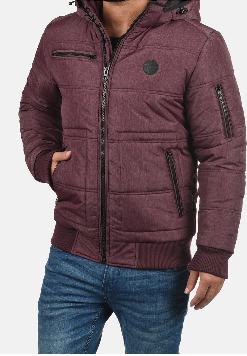 Bordeauxrote gesteppte Jacke mit Kapuze, ausgestattet mit Reißverschlusstaschen, einem gerippten Saum und einem runden Logo-Patch auf der Brust. Strukturierte Stoffoberfläche.