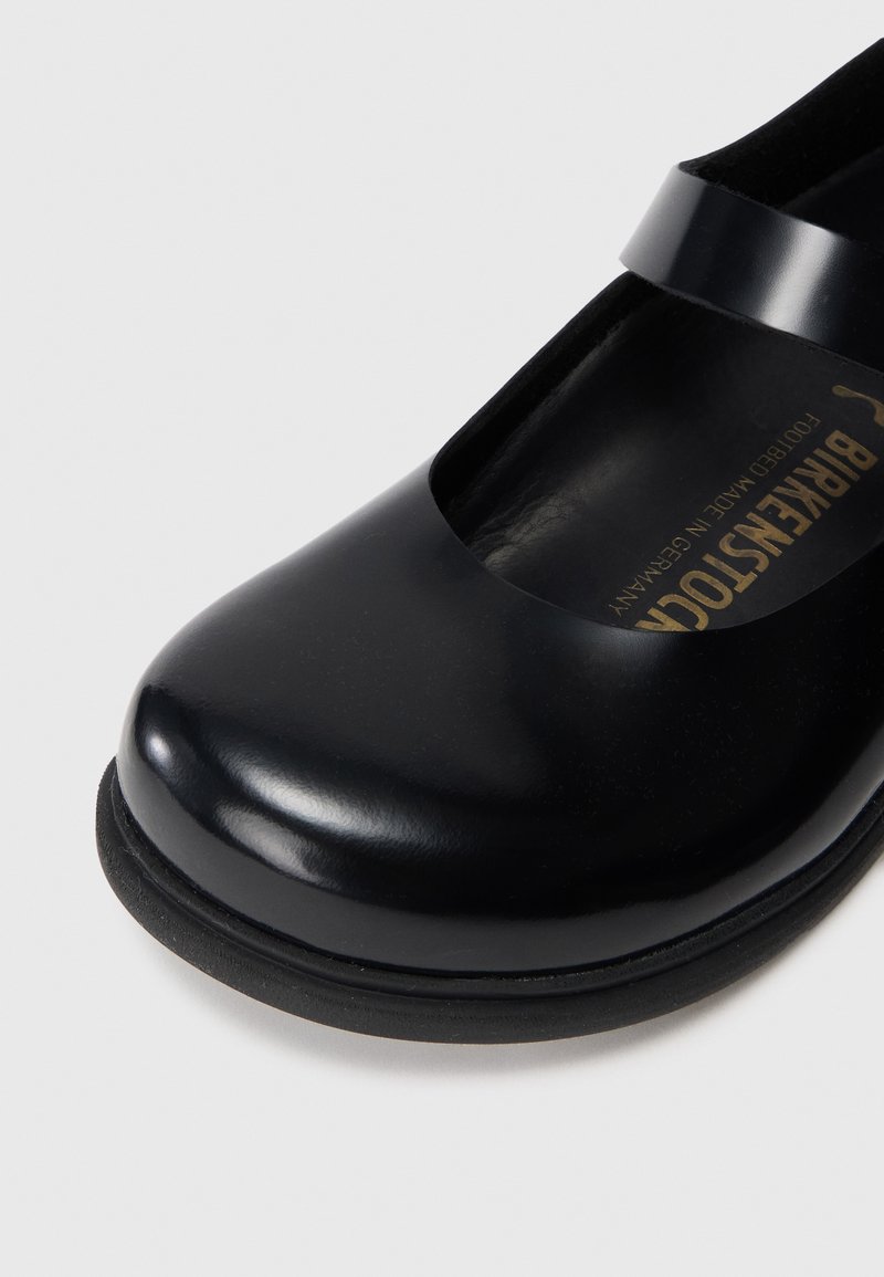 Zapato de charol negro con punta redondeada y una sola correa sobre el empeine. El interior presenta una marca dorada sobre un fondo oscuro.