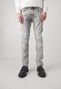 Jasnoszare, zwężane jeansy z wyblakłym wykończeniem i lekkim przetarciem, zestawione z czarnymi wsuwanymi butami, prezentowane na neutralnym tle.