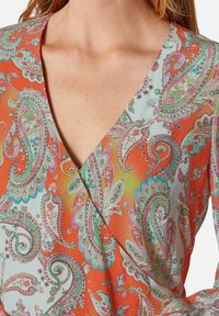 Orange gemustertes Kleidungsstück mit V-Ausschnitt und Paisley-Motiven in Grün und Pink, aus einem leichten Stoff gefertigt. Strukturierte Optik.