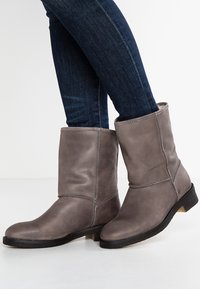 Šedé kotníkové boty z kůže s kulatou špičkou, prošitými detaily, měkkou texturou a černou gumovou podrážkou, nošené s tmavě modrými džínami.