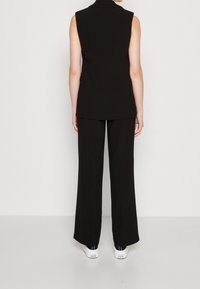 Schwarzes ärmelloses Oberteil mit hohem Kragen, kombiniert mit weiten schwarzen Hosen und weißen Sneakern. Glattes Material mit minimalen Details.