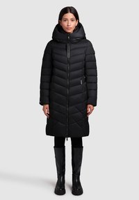 Black puffer coat with a quilted design, high collar, front zipper, and side pockets. Worn with black leggings and knee-high boots.