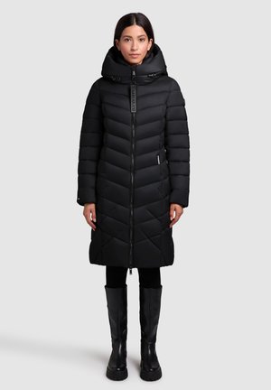 Zwarte pufferjack met een doorgestikt ontwerp, hoge kraag, rits aan de voorkant en zijzakken. Draagt samen met zwarte leggings en kniehoge laarzen.