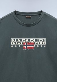 Tmavě zelené tričko s kulatým výstřihem, na kterém je bílé logo "NAPAPIJRI" a grafika norské vlajky, spolu s nápisem "geographic since 1987."