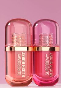 Zwei Flaschen flüssiger Rouge mit runden, durchsichtigen Kappen in Koralle und Pink, mit goldenen Akzenten und geprägten Etiketten.