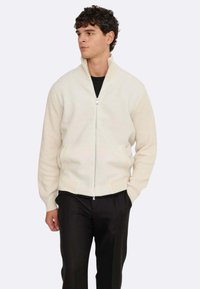 Cremfarbener Reißverschluss-Pullover mit ribbeliger Struktur, hochgeschlossen und mit kontrastierenden Strickmustern an den Ärmeln. Kombiniert mit schwarzen Hosen.