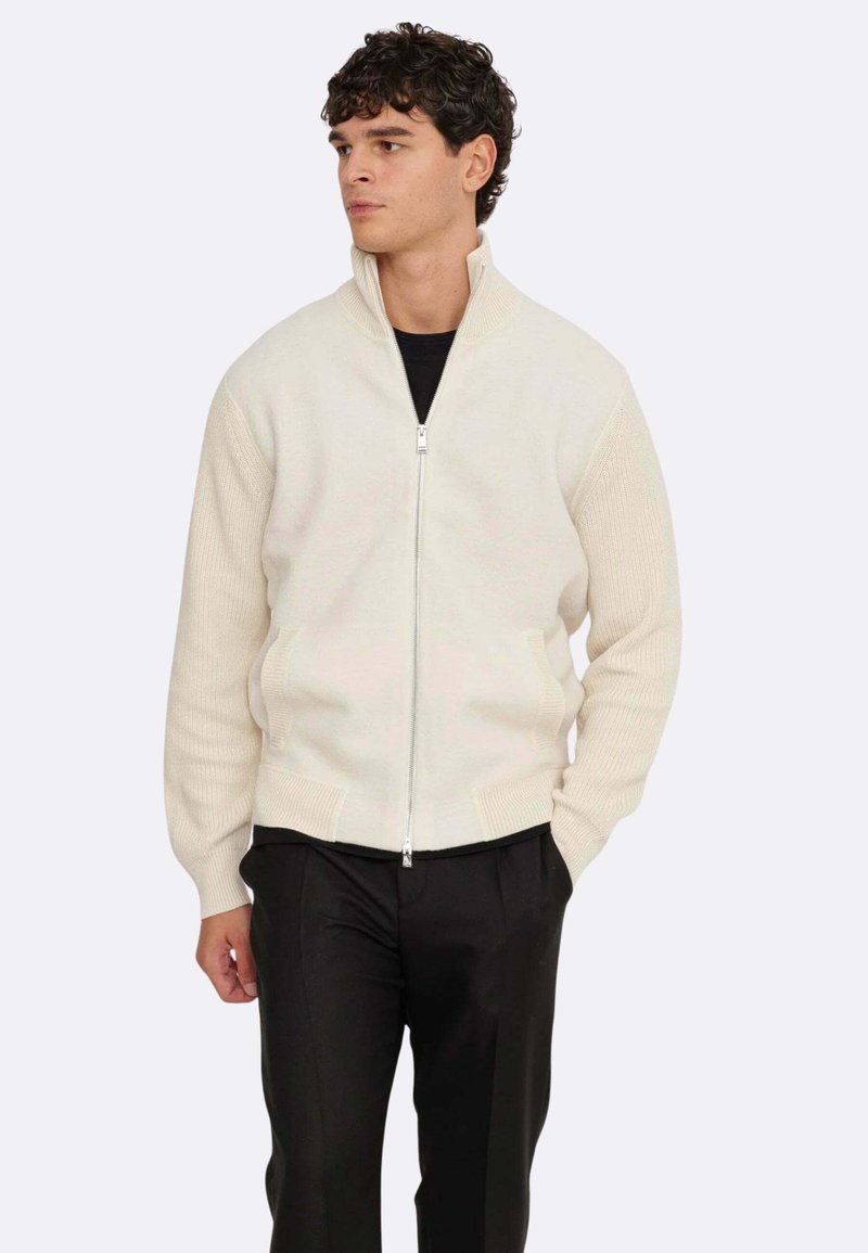 Cremfarbener Reißverschluss-Pullover mit ribbeliger Struktur, hochgeschlossen und mit kontrastierenden Strickmustern an den Ärmeln. Kombiniert mit schwarzen Hosen.