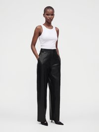 Femme portant un haut blanc sans manches, un pantalon large en cuir noir taille haute et des talons pointus noirs, debout les mains dans les poches.