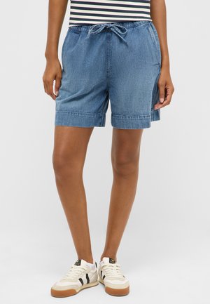 Person trägt blau mittelange Jeansshorts mit Kordelzug in der Taille, steht in weißen Turnschuhen mit schwarzen Streifen vor schlichtem Hintergrund.