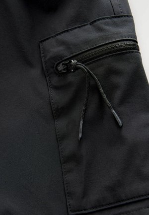 Črna tkanina z gladko teksturo, opremljena z žepom z zadrgo in detajlom s trakom za zategovanje na stranskem šivu. Čist in minimalističen dizajn.